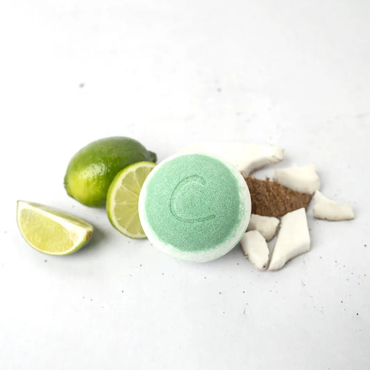 Round light green bath bomb with a letter C, surrounded by fresh lime wedges and coconut chunks on a white surface.