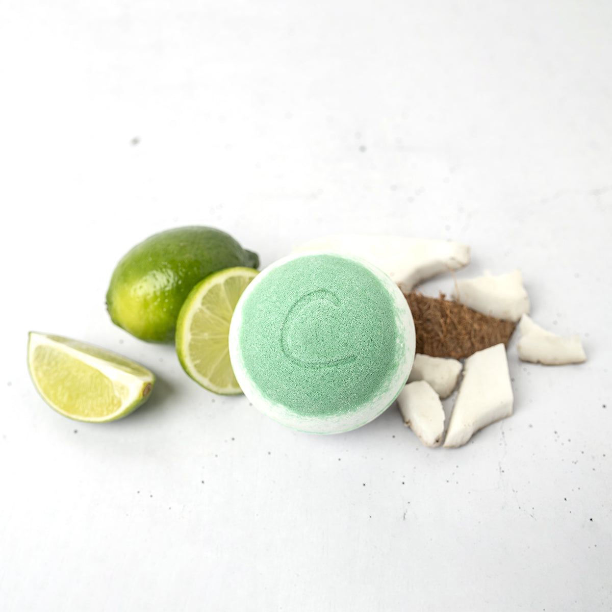 Round light green bath bomb with a letter C, surrounded by fresh lime wedges and coconut chunks on a white surface.