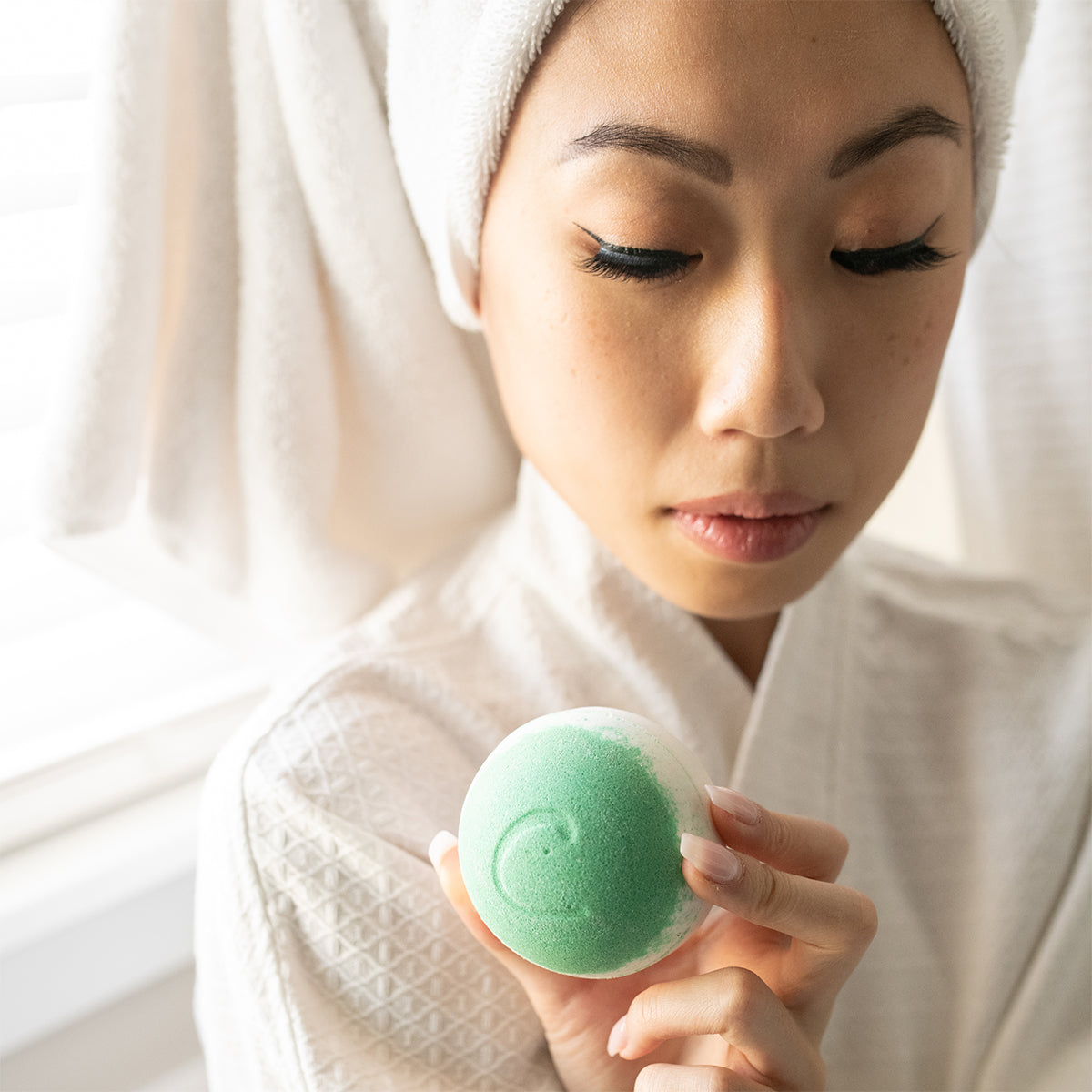 Woman in a white robe holding a round, green and white Coconuts About Lime moisturizing bath bomb.