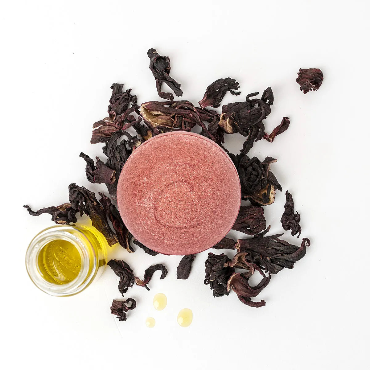Round pink bath bomb surrounded by dried hibiscus flowers and a small glass bottle of golden essential oil.