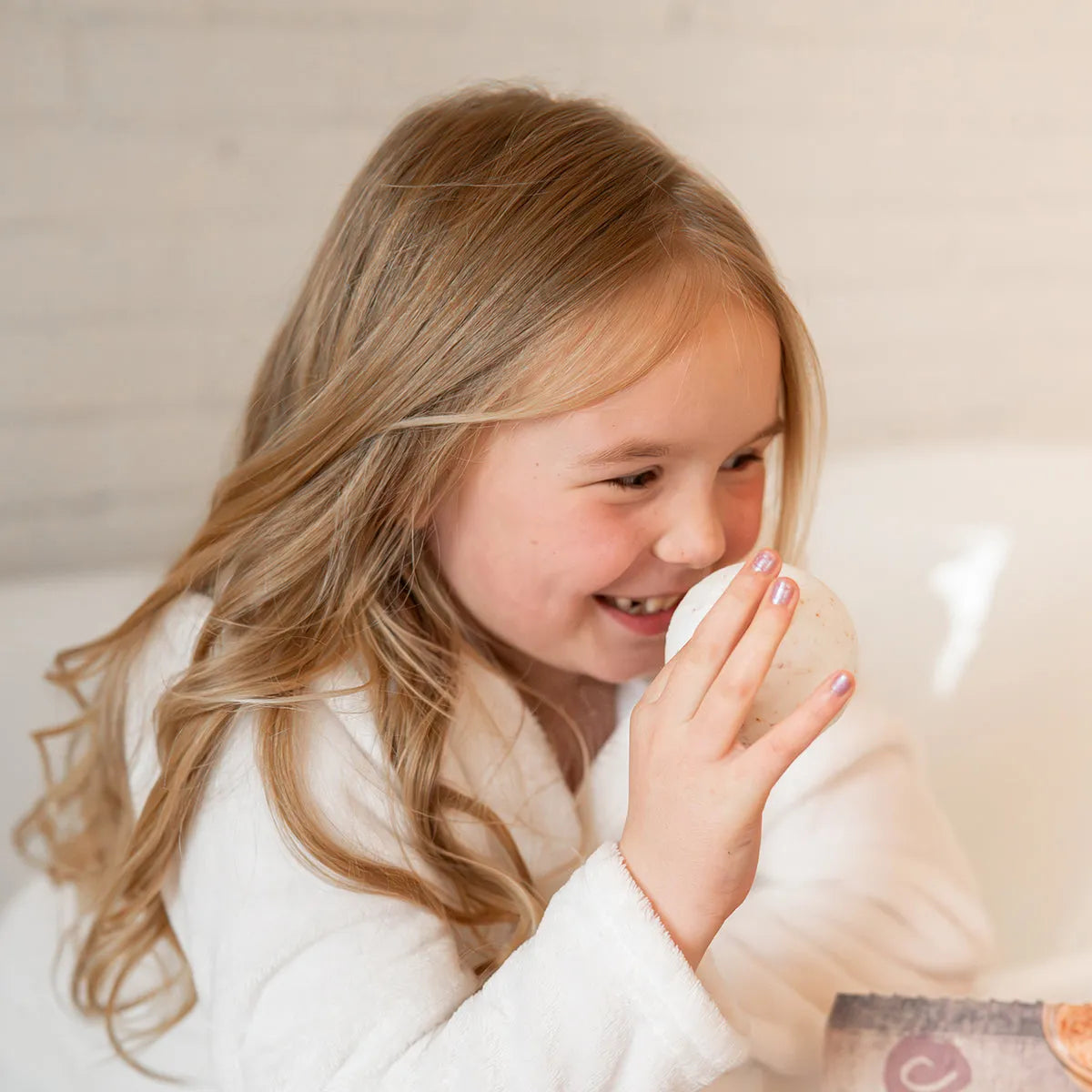 Young girl in a white robe smiling while holding a round, speckled Himalayan salt toning therapy bath bomb.