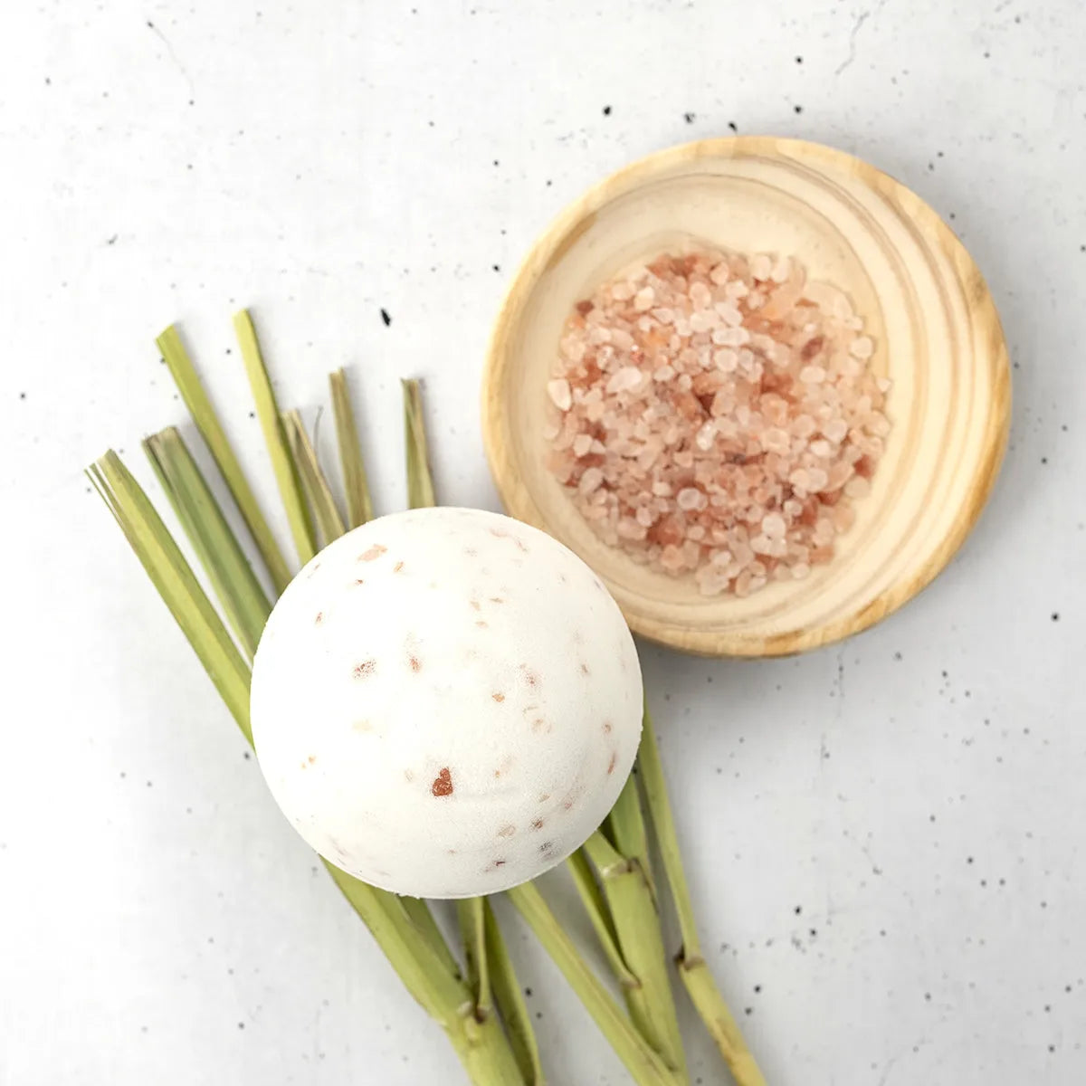 White Himalayan salt bath bomb with pink mineral flecks, next to a wooden bowl of salt and fresh lemongrass stalks.